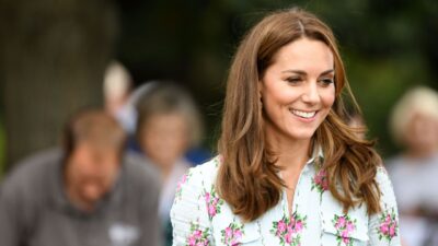 Kate Middleton, uma mulher branca de cabelo liso e castanho claro, aparece sorrindo. Ela está vestindo uma roupa branca, com golas, estampada com flores cor de rosa.